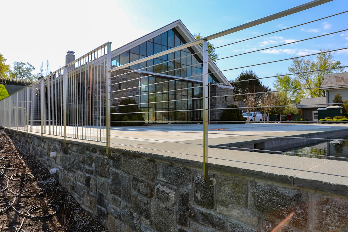 A cable railing in the foreground sits atop a stone retaining wall and connects to a metal fence in the background. Together the two block access to a pool on the right side of the photo.