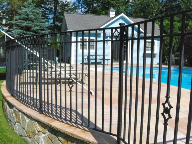 A wrought iron fence surrounds a small pool enclosure and adjacent blue pool house.