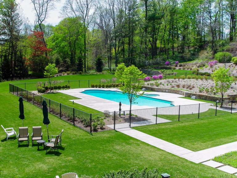 A bright blue pool nestles in a groomed landscape; it is surrounded by a black metal fence.