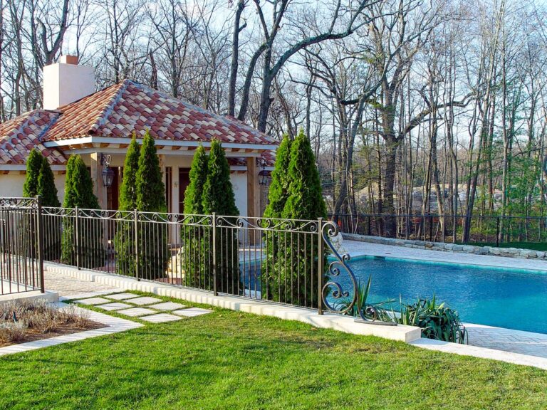 A wrought iron fence extends into frame between the viewer and a pool house with a tiled roof. The fence has vertical bars and is topped with decorative scrollwork; an ornate decorative element protrudes from the final fence panel.