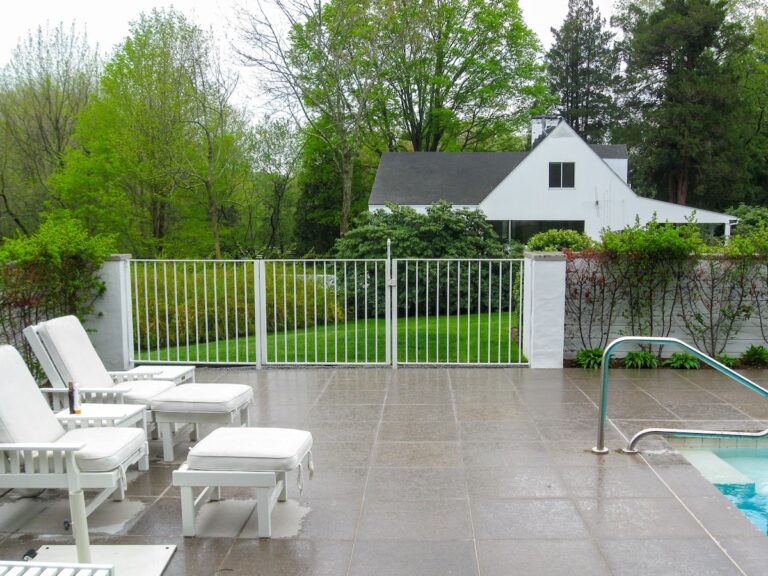 A three-paneled white stainless steel gate sits in between stone walls; white lounge chairs sit on large gray concrete tiles.