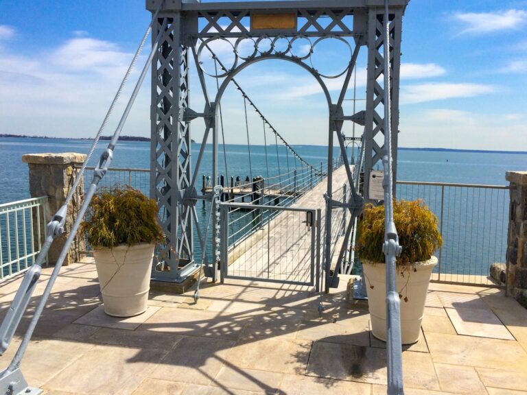A metal fence gate under a complicated custom-made archway designed to look like the George Washington Bridge leads out onto a long pier that extends out into the water.