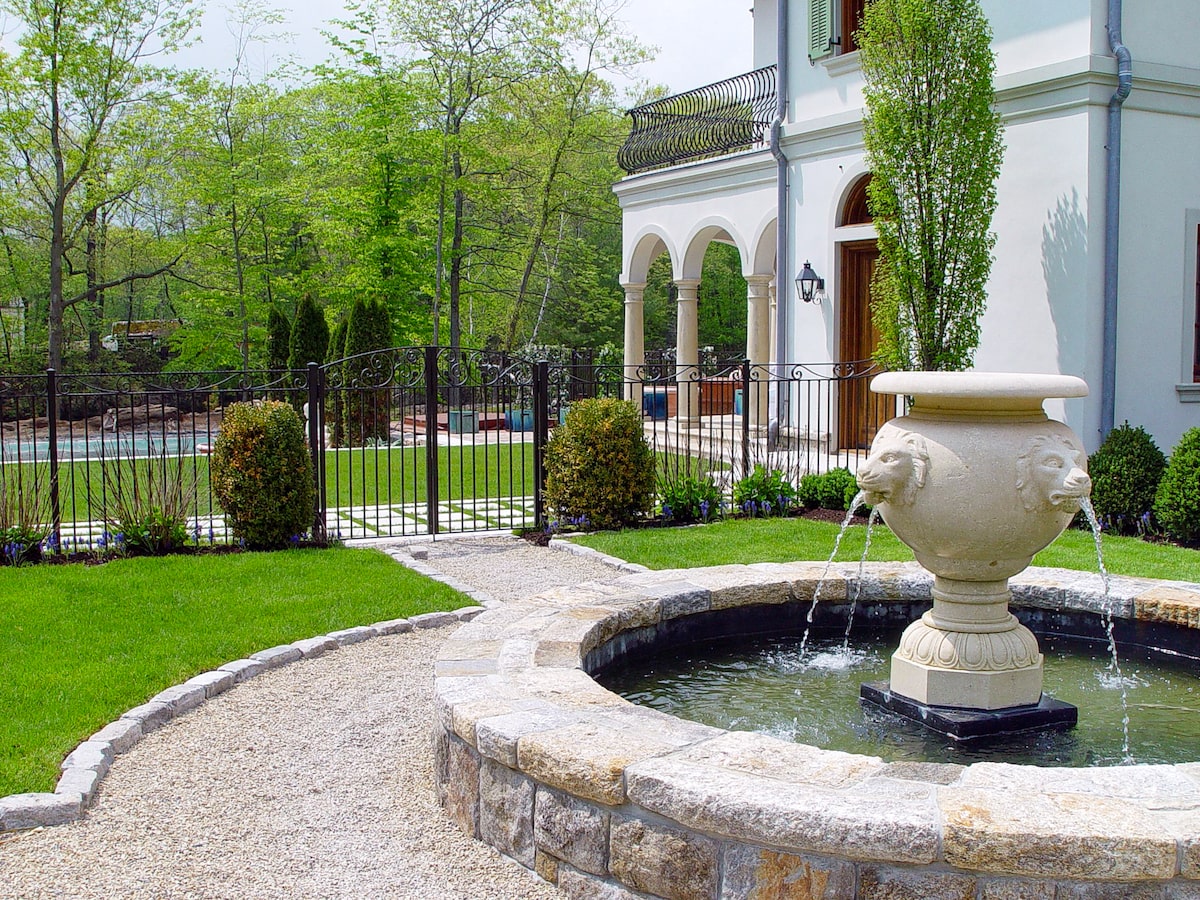 A dark-colored metal fence gate made of iron with ornamental scrollwork leads from a home’s outer yard to a fenced-in courtyard with a fountain in the foreground.