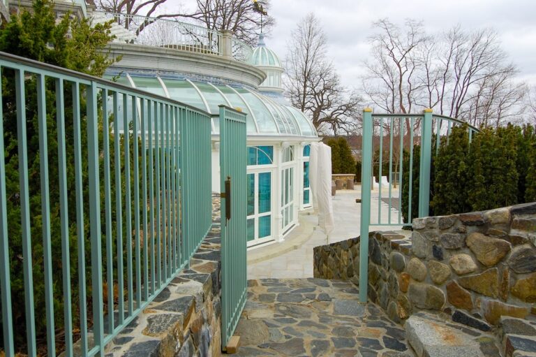 A green iron fence gate with a bronze top rail leads from a stone walkway to an ornate glass-domed indoor porch.