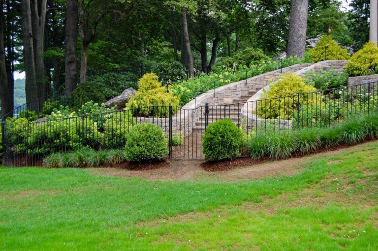 A simple metal fence gate leads to a stone staircase surrounded by lush landscaping. The gate is set in an iron fence that follows the gradient of the hill.