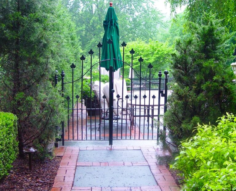 A metal fence gate with an arched top rail and finials at the top of its pickets leads to a fenced-in pool area. There are bushes on either side.