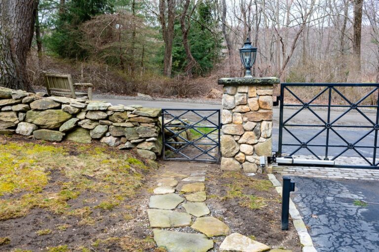A metal walk gate allows foot traffic along a slate path that leads from the main roadway through a low stone wall to the property within. The design of the walk gate matches that of the driveway gate right next to it.