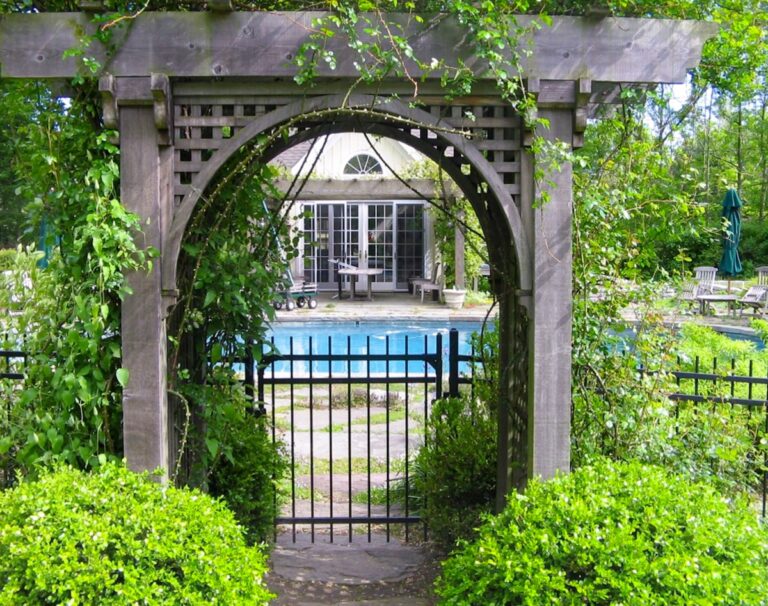Underneath a wooden arbor entrance covered with climbing vines, there’s a simple metal fence gate leading to a pool in the background.