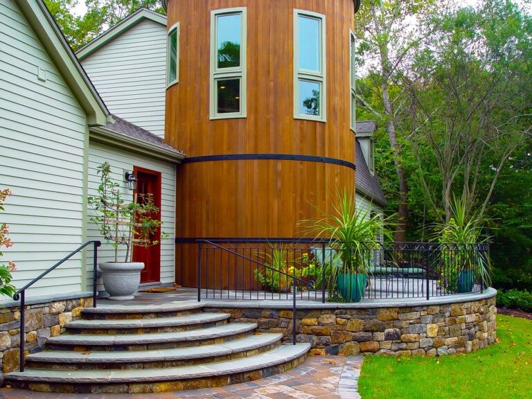 Minimal outdoor metal handrails in a V shape frame a set of semi-circular stone steps. There is a circular porch that goes around a wooden tower which is also circular.