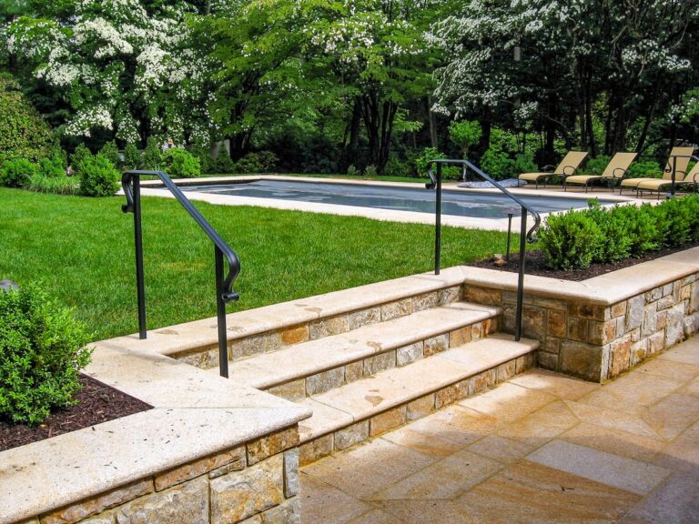 Three stone steps with a simple outdoor metal stair railing made from wrought iron lead from a stone patio to a grassy area. Behind the grass is a small in-ground pool.