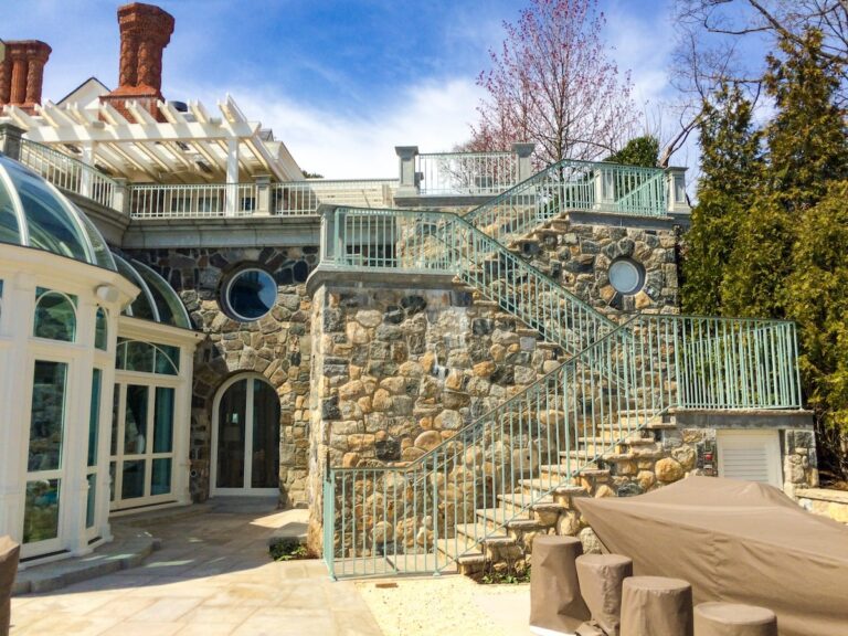 Green-painted wrought iron and bronze outdoor stair railings run alongside stone steps that go up two stories from a stone patio to an observation deck. There is a circular glass-walled sunroom next to the steps.