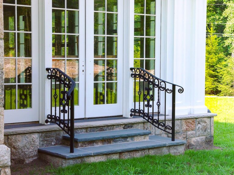 Ornate metal railings sit alongside two short stone steps that lead from a grassy lawn to white French doors.