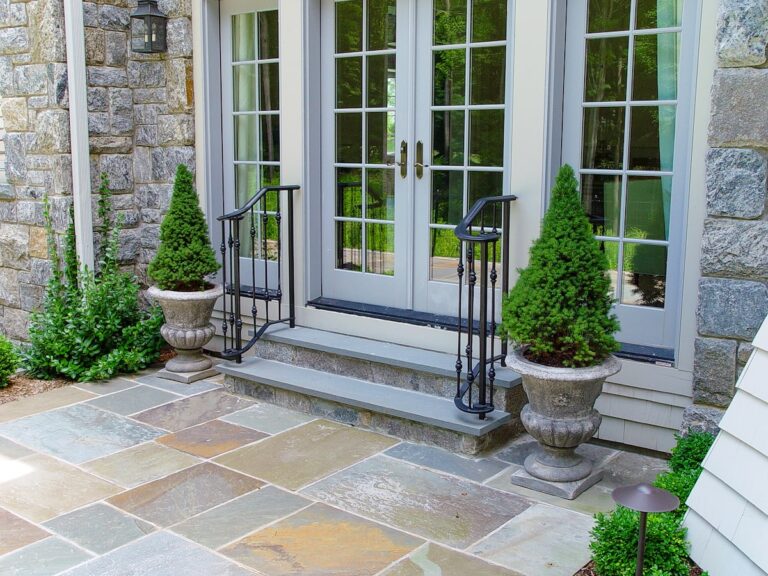 A flagstone patio of different stones is in front of a pair of white French doors. Leading between the doors and the patio are two stone steps with a simple wrought iron stair railing that curves at the base step.