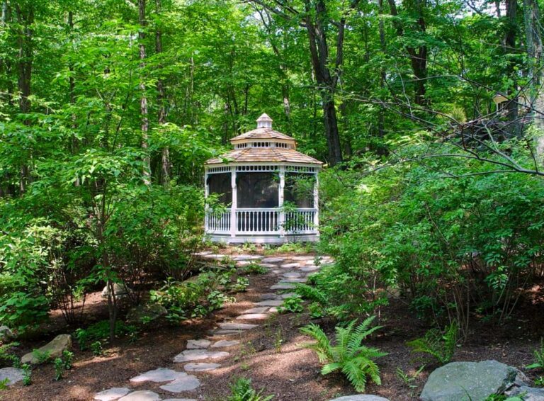 A gazebo made from white-painted wood lies at the end of a stone footpath through the woods.