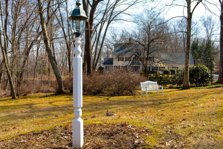 A white wooden post with decorative details supports a black metal lantern on top. A bench and a home can be seen in the background.