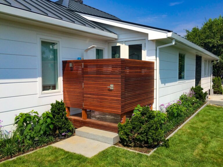 A mahogany shower enclosure made from tightly spaced thin strips of wood fits into a nook along the exterior sidewall of a home.