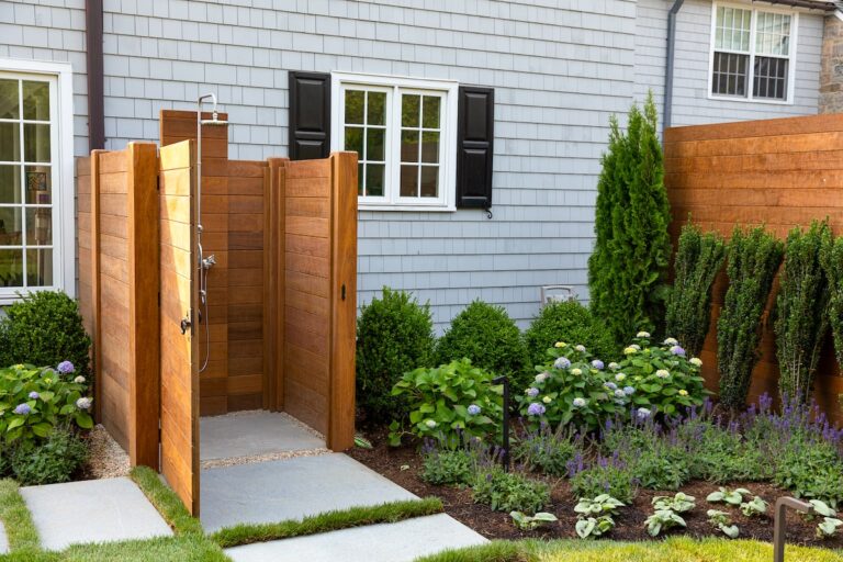 A spacious outdoor shower enclosure made from solid-paneled dark-stained wood is placed by the side of a home.
