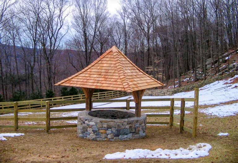 A decorative dry-fit stone well with a wooden roof sits next to a wooden fence on a wide open piece of property surrounded by forested hills.