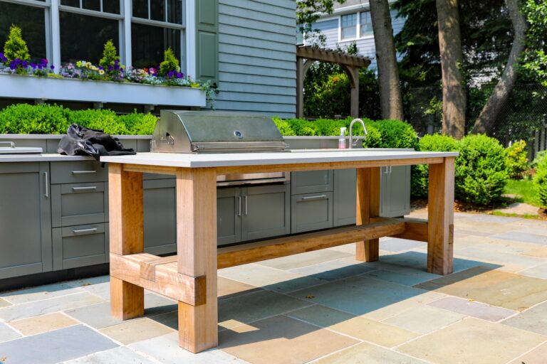 A custom made kitchen island rests on the flagstone floor of an outdoor kitchen behind a home. Behind it is a grill and a sink built into outdoor cabinets.
