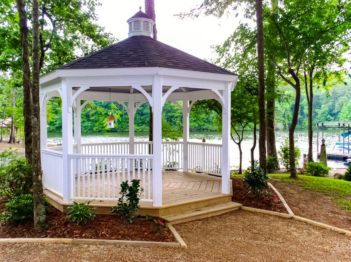 A classic white wood gazebo with a black shingle roof sits beside the water. A little birdhouse hangs down in the middle of it.