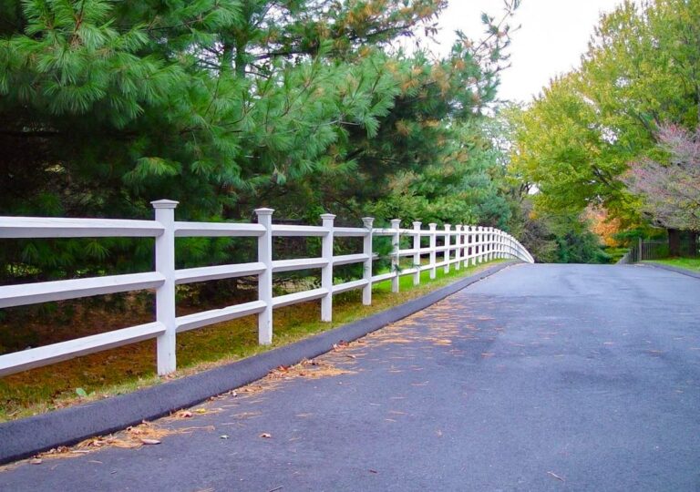 A simple white post and rail fence with 3 rows of rails borders an asphalt drive on the right and tall evergreen trees on the left.