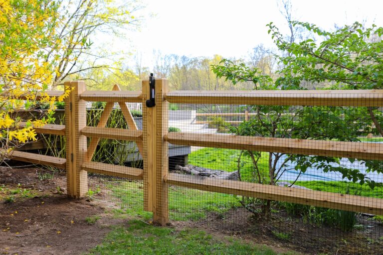 A swing gate interrupts the run of a wood post and rail fence. The fence and gate are covered with wire mesh to meet local building codes for the pool enclosed within.