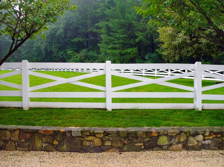 A white post and rail fence surrounds a horse paddock. The rails are shaped in an x pattern with a double bottom horizontal rail.