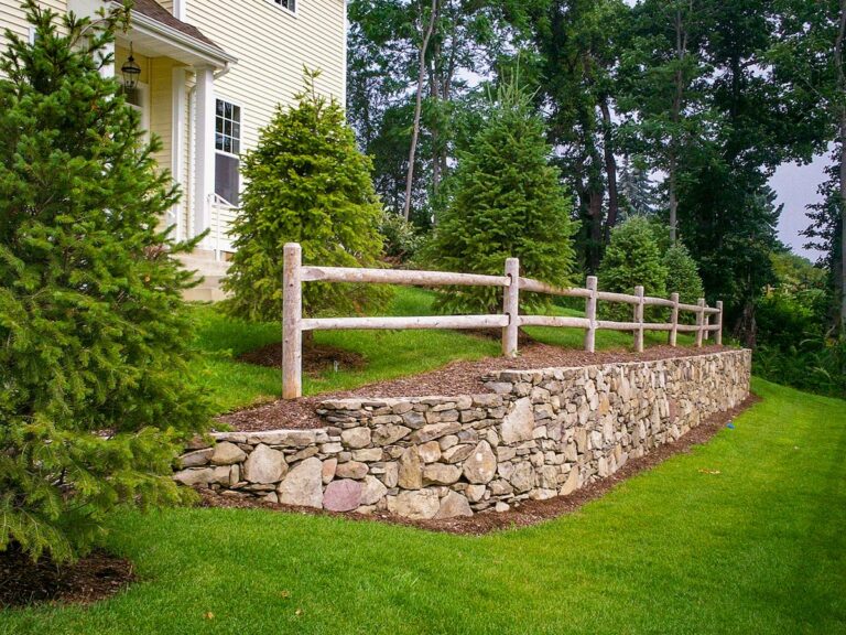 Wooden post and rail fencing runs along the top of a stone retaining wall next to neatly landscaped evergreen trees and a lawn sloping down from a house.