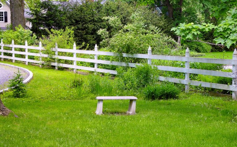 A small stone bench sits on a lawn in front of a white post and rail fence.