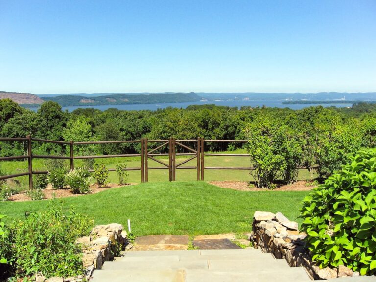 The camera looks out towards water in the far distance, over a tall wooden post and rail fence in the foreground and a forest in the middle distance. Stone steps lead down towards the fence.