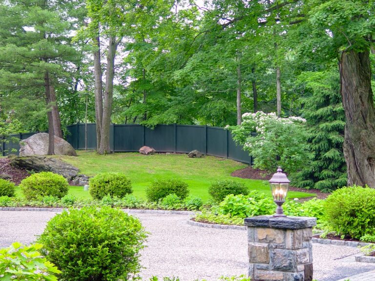 A dark green privacy fence blends into the landscape; a shrub blooms white in the foreground.