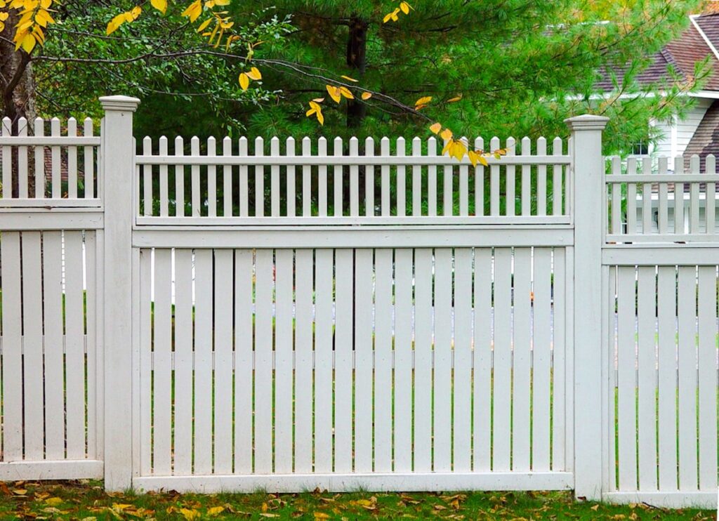 A white picket fence with the pickets set close together but not touching blocks most of the view of a yard.