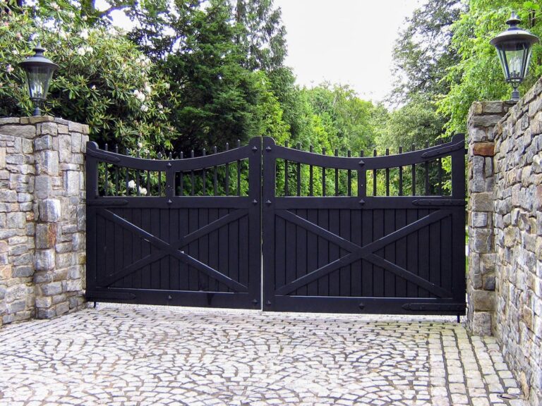 A black paneled gate hangs between two stone pillars. The road is paved with stone as well.