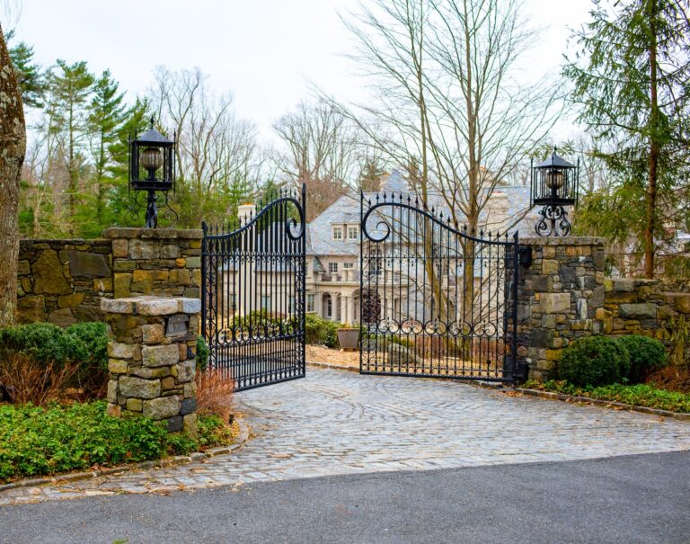 Two panels of a wrought iron gate protect a stone drive leading to an imposing house in the background. The gate is hung between two rustic stone pillars topped with wrought iron lamps.