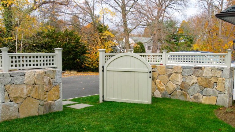 A low stone wall is topped with a narrow strip of white lattice fencing. A half gate in the wall stands open between a driveway and a yard.