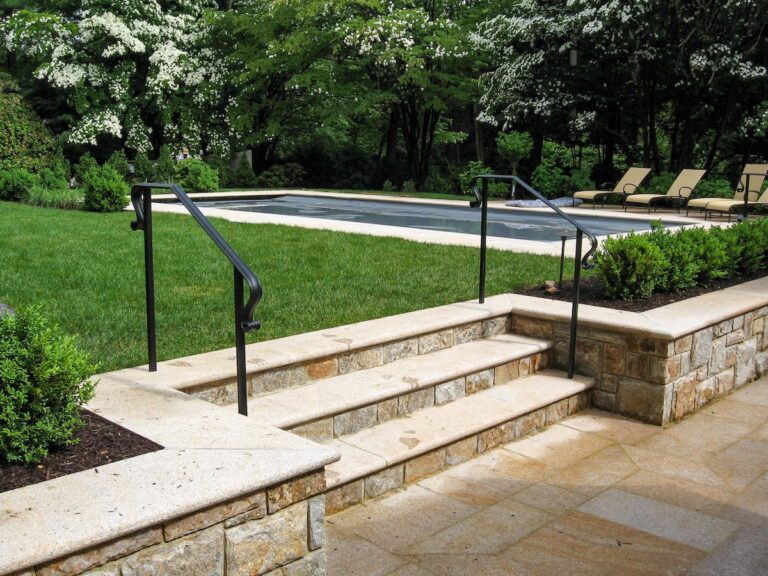 Three stone steps lead from a sunken paved area to the grass surrounding it. There’s a simple iron handrail on either side of the stairs.