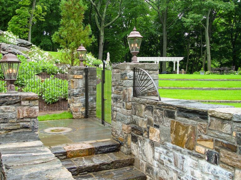 Steps and a stone walkway lead between two stone half-walls to a wrought iron walk gate flanked by stone columns topped with lanterns.