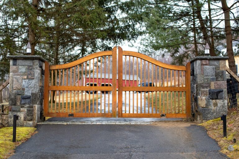 A wooden gate with vertical rails sits between stone posts. There is a red barn in the background.