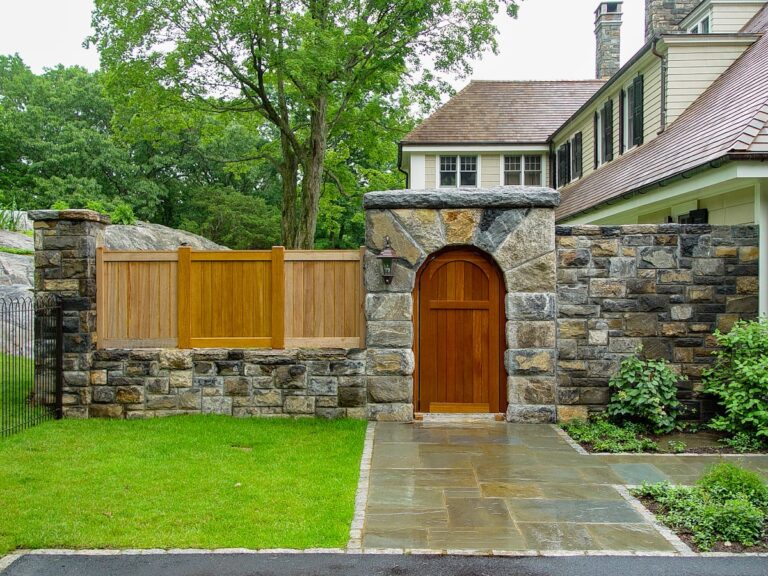 An arched wooden door is set into a stone wall. A flagstone path leads to the door, and a wooden privacy fence shields the yard from view.