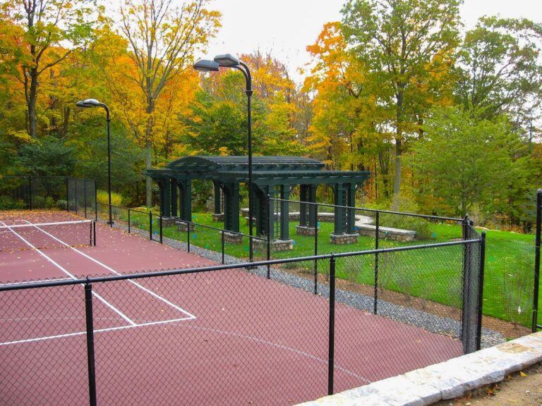 The viewer sees the corner of a tall chain link fence that surrounds a dark red tennis court. There is a large wooden pergola beyond the court.