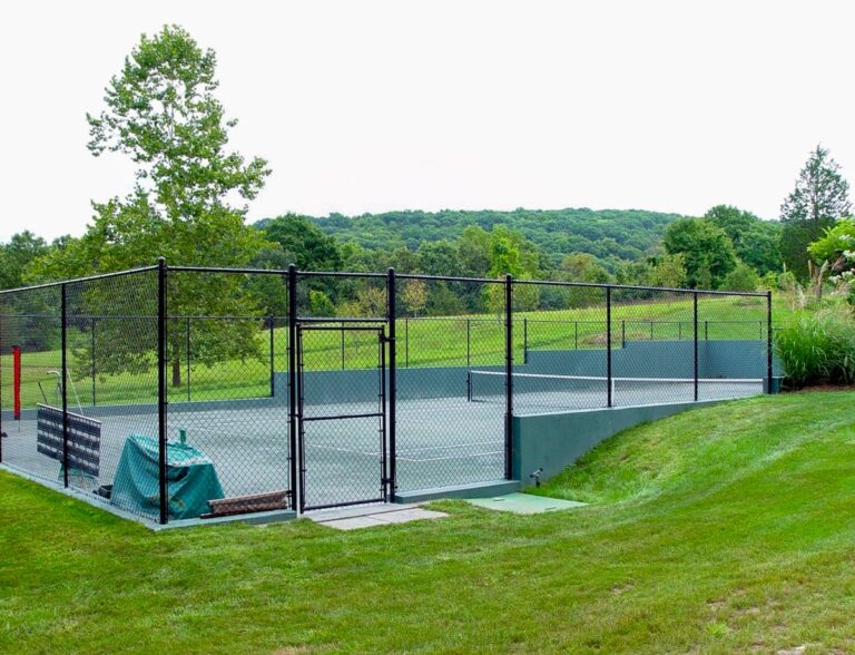 A tall black chain link fence surrounds a tennis court that is set into a gentle slope in a green lawn.