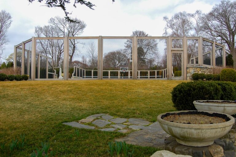 The viewer looks across a lawn towards a tall wood and metal mesh tennis court fence.