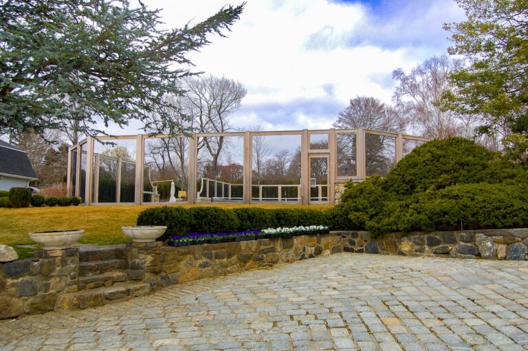 The viewer looks up from a sunken stone patio toward a tall wood and mesh fence surrounding a tennis court.