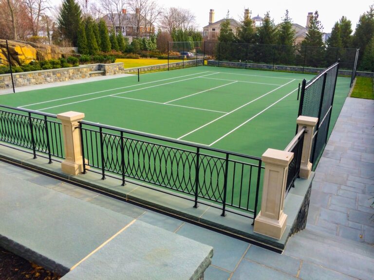 A short decorative metal fence surrounds a green tennis court. The court is set within a flagstone patio, which is bounded by a small stone wall.