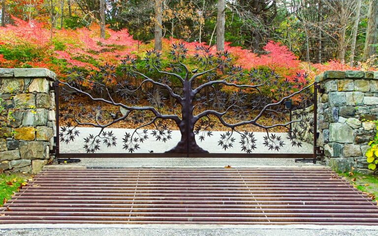 A custom wrought-iron double gate sits between 2 dry fit stone pillars. The gate is in the shape of a tree, with branches and leaves meeting at the center trunk. The trunk splits to swing open.