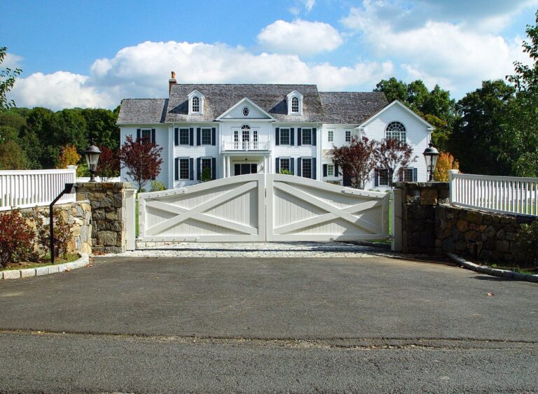 Two stone walls curve in to meet a solid white wood double gate in front of a large white Cape home.