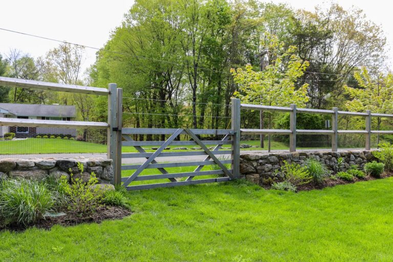 A wide wood fence gate with diagonal cross beams sits between a low stone wall with a tall split rail fence built on top.