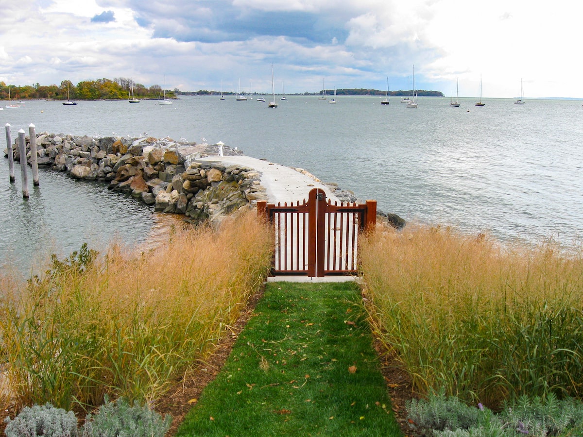 A dark-stained wood walk gate with metal trim leads out to a path along a stone jetty overlooking the water.