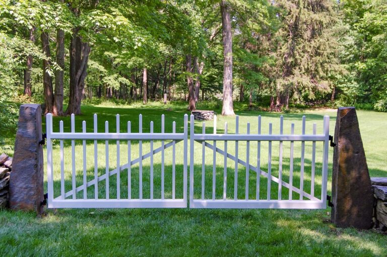 A long, wide white wood fence gate between two stone columns leads to an open lawn with woods in the background.