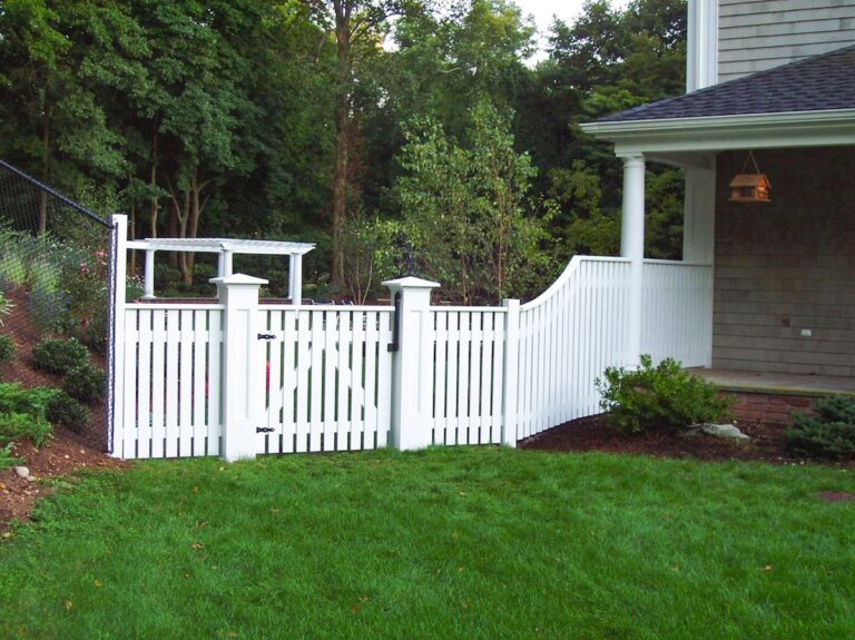 A white picket fence gate leads from the lawn in the backyard to a more open space on the side of the house with a matching white pergola in the background.